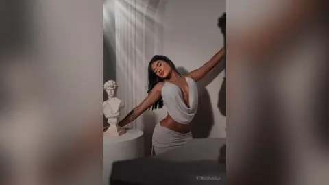 Elegant woman in a white plunging maxi dress posing by a classical bust in a minimalist, sunlit studio.