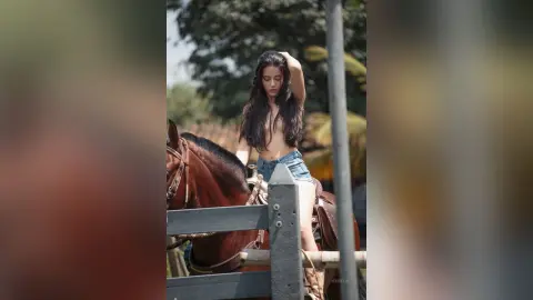 JessieLiu sits naked on a brown horse, running her hand through her long dark hair outdoors.
