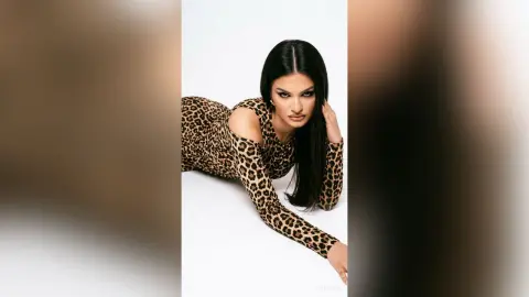 ZenyaSin poses lying down in a long-sleeved leopard print bodysuit against a white background.