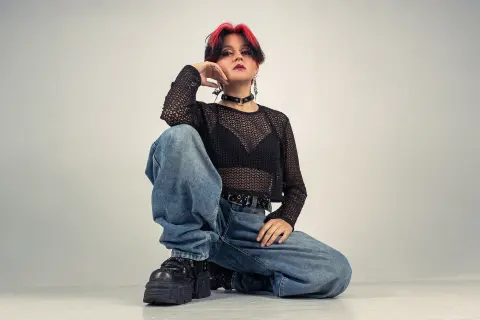 YuuMiko poses kneeling in baggy blue jeans and a black mesh long-sleeve top, showing her red-tipped hair and dark choker against a plain white background.