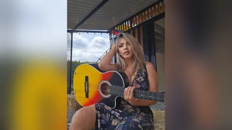 CrisstyBaez — CrisstyBaez sits outdoors holding an acoustic guitar while wearing a floral print sundress.