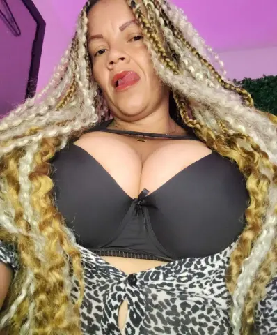 Woman with long blonde braids wearing a black bra and leopard print top in a soft pink lit room.