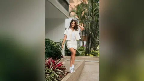TefyRusso smiles while posing outdoors in a white short-sleeved jacket and matching mini skirt, standing near green tropical plants.