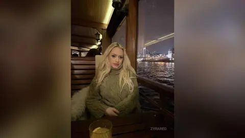 Blonde woman in a cozy green knit sweater sitting on a boat at night with city lights reflecting on the water.