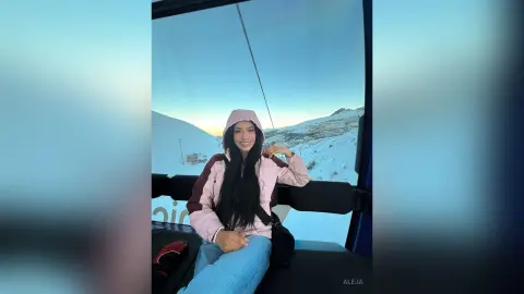 Aleja smiles while sitting in a ski lift wearing a pink hooded jacket and blue jeans against a snowy mountain backdrop.