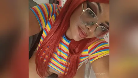 Smiling woman with long red hair wearing a colorful striped crop top in a bright indoor setting.