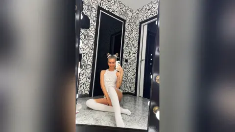 Smiling woman in a white lace mini dress and thigh-high boots posing in a modern black and white patterned room.