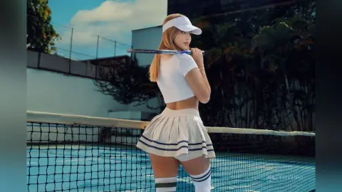 Blonde woman in white tennis outfit holding a racket on a sunny outdoor tennis court.