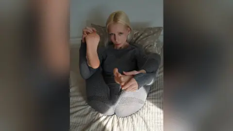 Blonde woman in a grey loungewear set sitting cross-legged on a bed in a cozy, relaxed indoor setting.