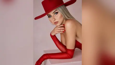Elegant blonde woman in a red wide-brimmed hat and matching satin gloves posing against a soft pink background.