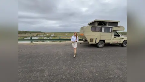 Woman in white shirt and shorts standing by a tan camper van on a bridge under a cloudy sky.