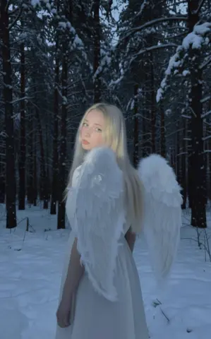 Blonde woman in a white ethereal angel costume with large feathered wings in a snowy winter forest.