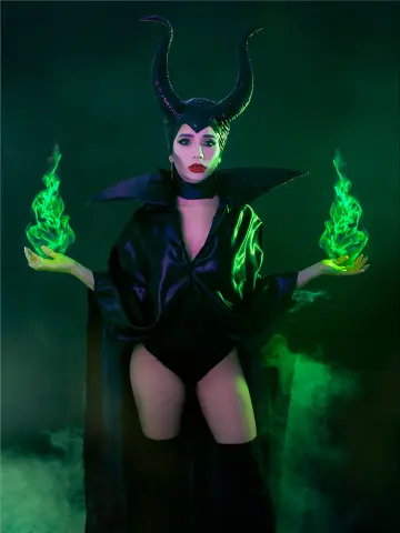 Woman in Maleficent cosplay with black horns and flowing cape holding glowing green magical flames in a misty setting.