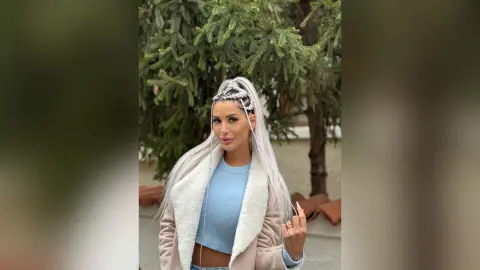 Woman with silver braided hair wearing a light blue sweater and beige fleece jacket in a lush garden setting.