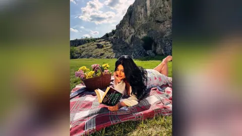 Young woman in a floral dress reading a book on a picnic blanket in a sunny meadow near rocky cliffs.
