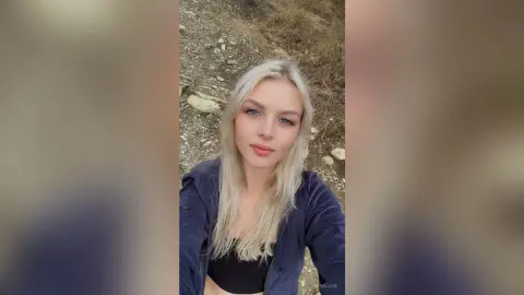Blonde woman in a black top and navy jacket smiling outdoors in a natural rocky landscape.