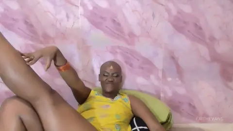 Smiling woman in a yellow patterned top relaxing against a pink marble wall in a bright, casual setting.