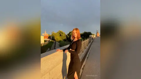 Young woman in a black long-sleeve dress standing on a stone bridge during a warm sunset.
