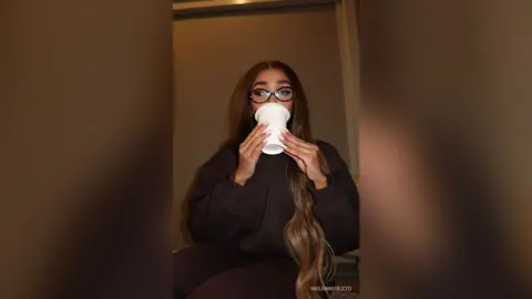 Young woman with long hair and glasses sipping coffee in a cozy, moody indoor setting.