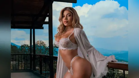 Blonde woman in white lace lingerie and robe posing on a balcony overlooking a scenic mountain landscape.
