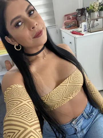 Young woman in a gold patterned crop top and denim jeans posing in a bright room.