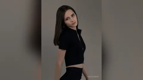 Young woman in a chic black zip-up crop top posing against a minimalist grey background with a moody expression.