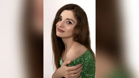 Smiling young woman with long brown hair wearing a green floral off-the-shoulder dress against a white background.