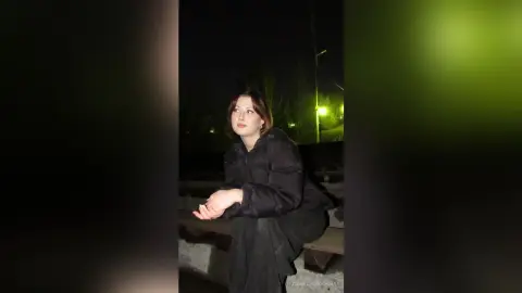 Young woman in a black puffer jacket sitting on a park bench at night under dim streetlights.