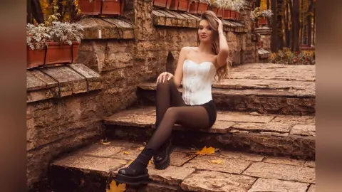 Young woman in a white corset and black tights sitting on stone steps in a warm autumn garden setting.