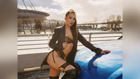 DelanaGrace poses leaning against a blue car wearing a black leather jacket and black lace lingerie with gold chain details.