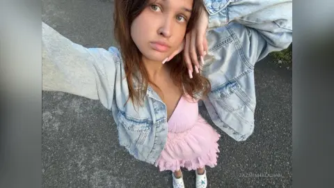 JazminHalajian wearing a light pink ruffled dress and a light wash denim jacket, posing outdoors with her hands near her face.