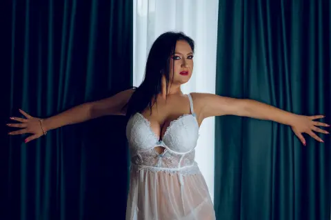 Woman in white lace lingerie posing with arms outstretched against dark green curtains in a moody setting.