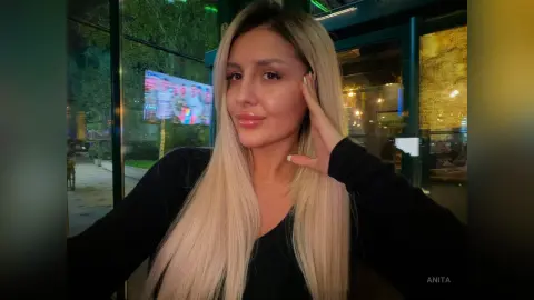 Anita poses with one hand against her cheek, wearing a black long-sleeved top with her long blonde hair falling over her shoulders.