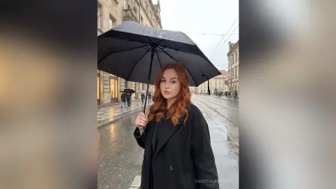 Redheaded woman in a black coat holding a black umbrella on a rainy city street.