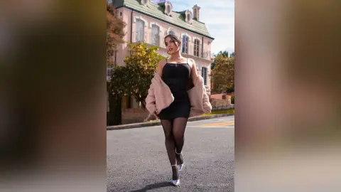 Stylish woman in a black mini dress and pink faux fur coat walking on a street near a classic building.