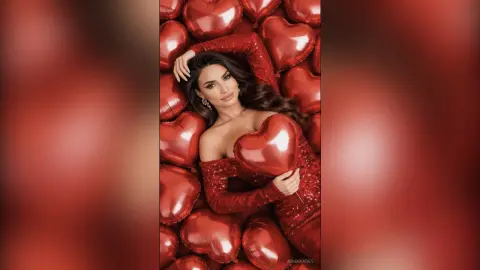 AnnaKarev poses with long dark hair and a red dress, holding a large red heart balloon against a background of red heart balloons.
