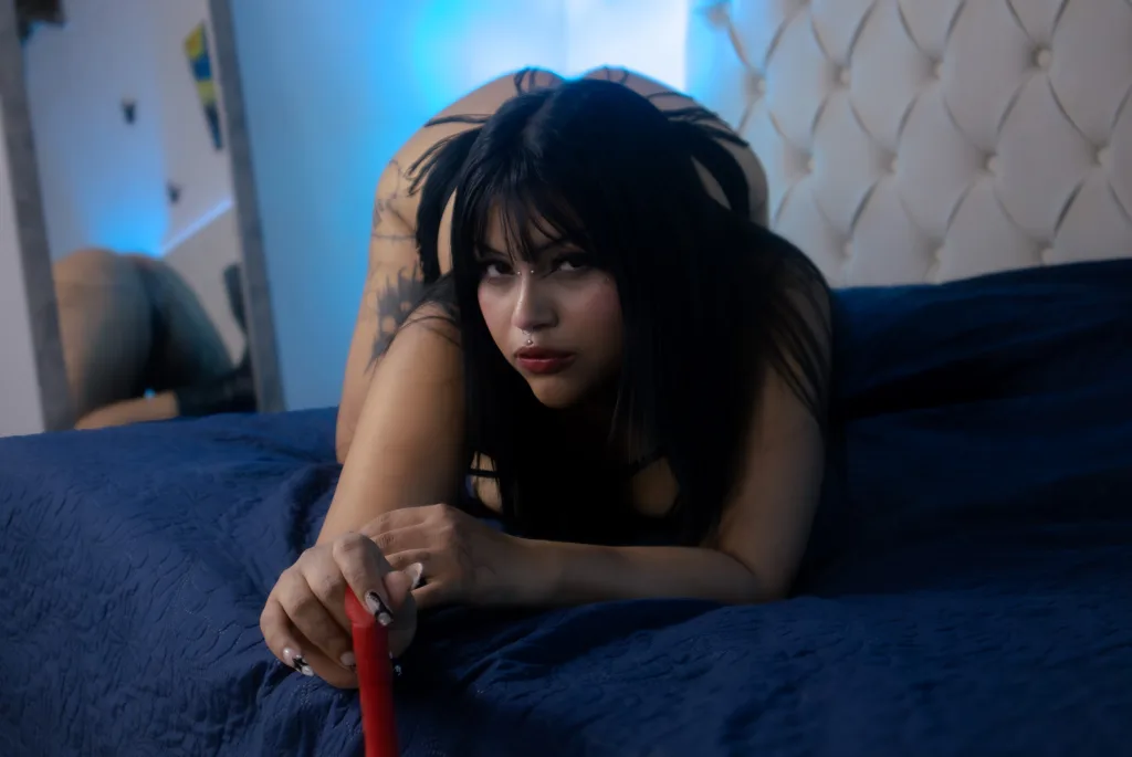 AngelicCopal poses with a direct gaze, wearing a black bra and black gloves against a dark red background.