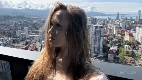 Young woman in a white top looking away on a city rooftop balcony overlooking a sprawling urban skyline.
