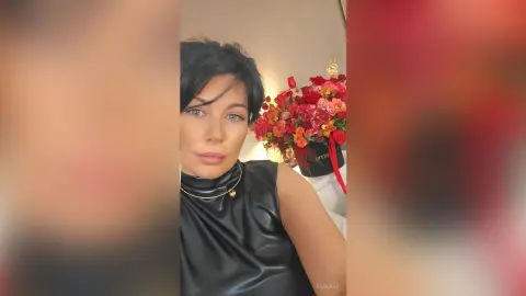 Woman in a black sleeveless top posing with a bouquet of red roses in a bright indoor setting.