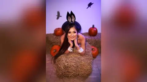 Woman in bunny ears costume leaning on hay bales surrounded by pumpkins in a spooky Halloween setting.