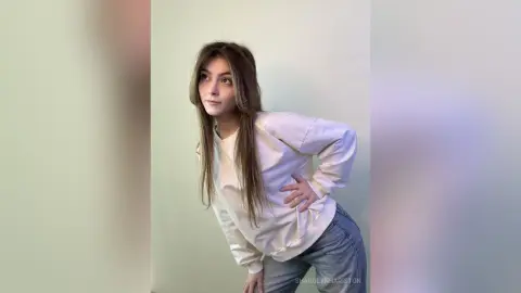 Young woman in a white long-sleeve shirt and blue jeans posing thoughtfully against a plain light background.