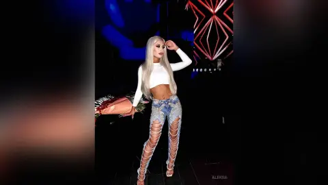 Glamorous blonde woman in a white crop top and ripped denim jeans posing at a night event with flowers.