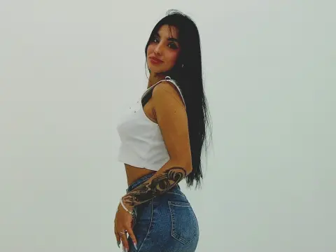 Ema_Velvet poses sideways in a white crop top and denim jeans, showing off her long dark hair and arm tattoo against a plain white background.