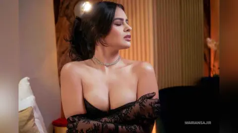 Elegant woman in a black lace outfit and silver necklace posing gracefully in a warm, moody indoor setting.
