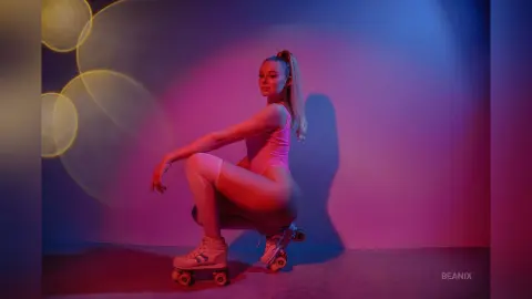 BeaNix squats in a pink bodysuit and white socks, posing against a blue and red lit wall.