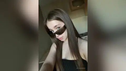Young woman with long brown hair wearing dark sunglasses and a black tank top in a bright indoor setting.