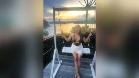 Woman in a black bikini and white sarong sitting on a swing overlooking a tropical ocean sunset.