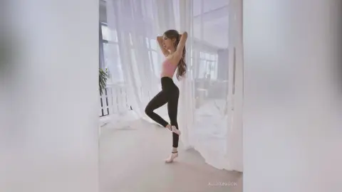 Fit woman in pink sports bra and black leggings posing gracefully in a bright, airy room with white curtains.