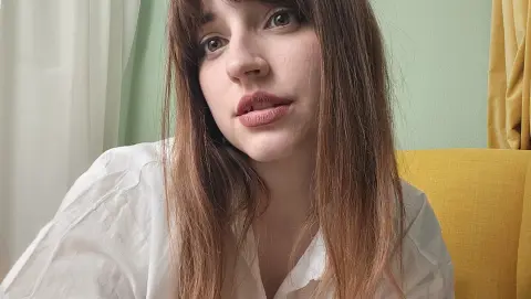Young woman with long brown hair wearing a white linen shirt in a bright, cozy indoor setting.