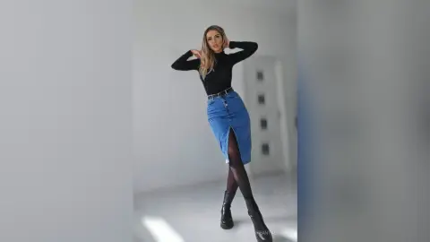 TiffanyAddams poses in a black long-sleeve top and a blue denim slit skirt with black tights and black boots against a plain white background.